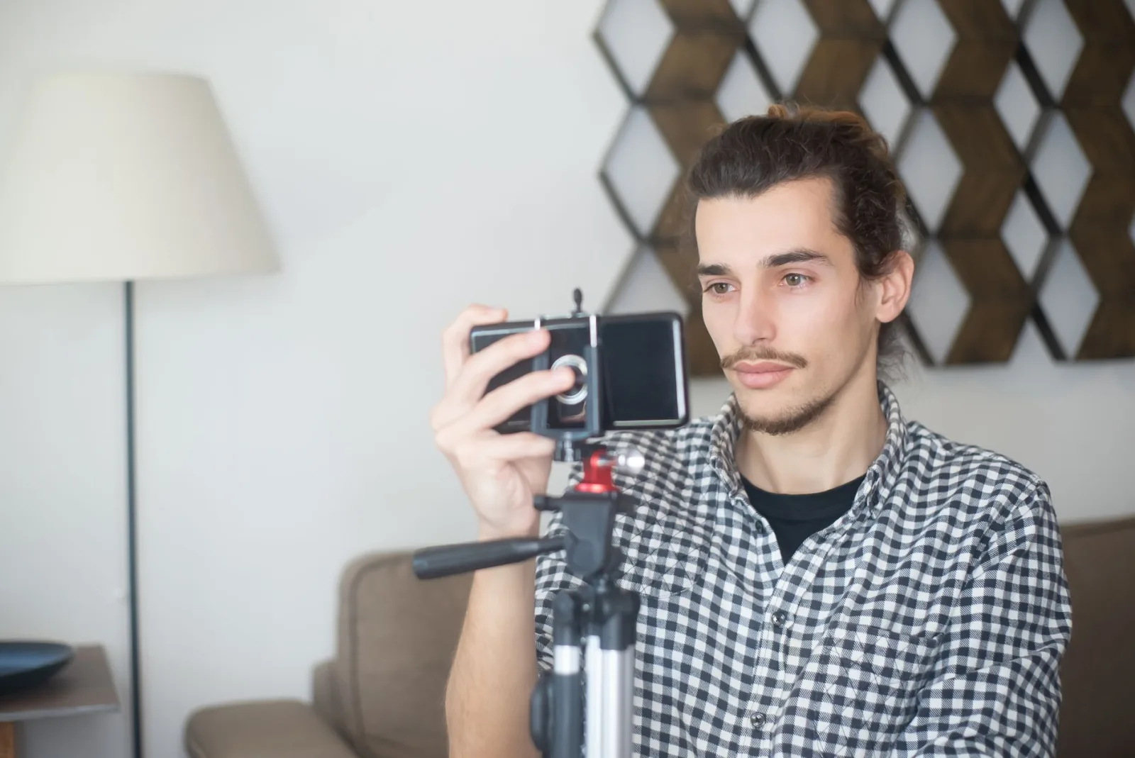 A man with a moustache and tied-back hair frames a shot on a smartphone mounted on a tripod in a living room