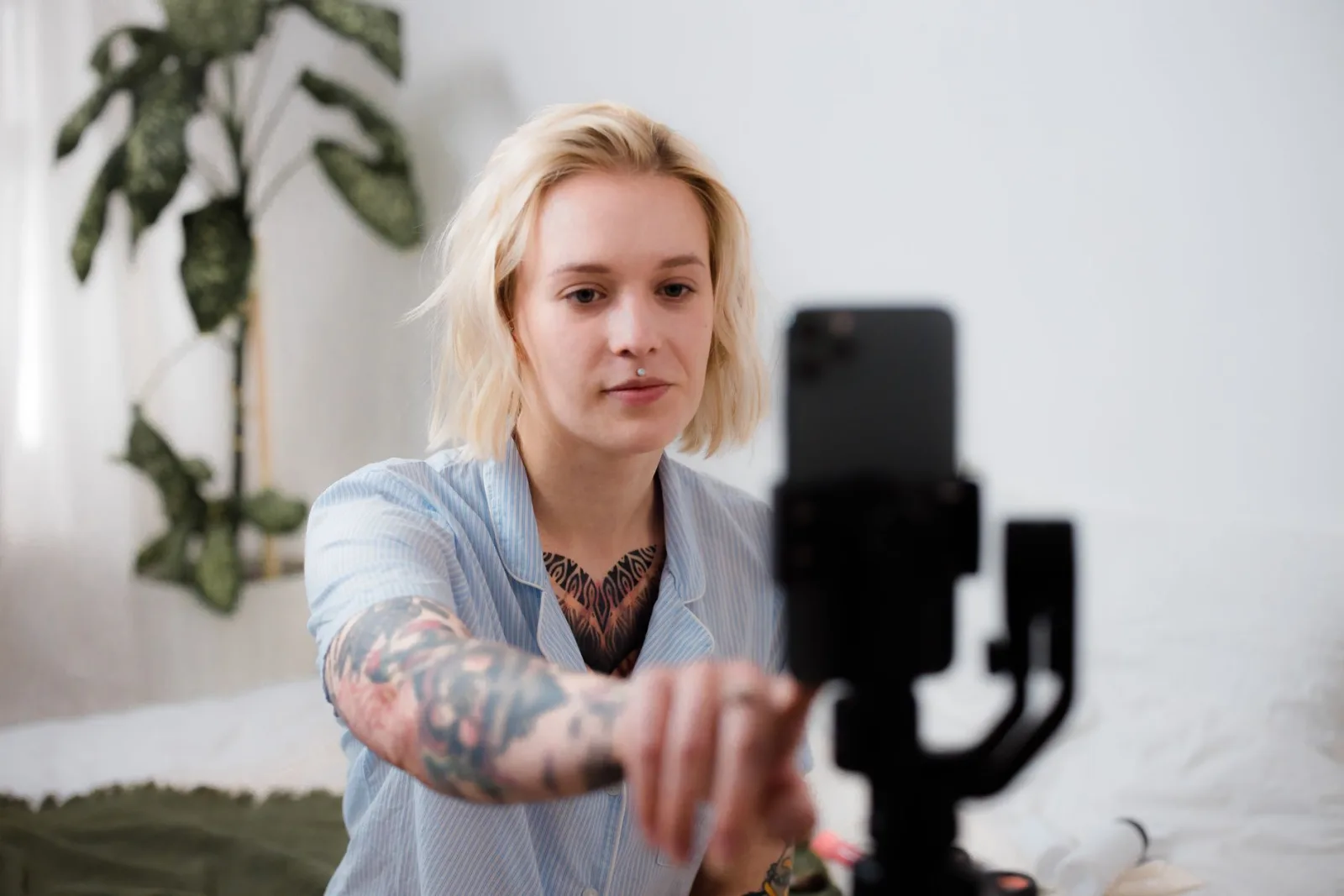 A blonde-haired creator with tattooed arms reaching to adjust an iPhone mounted on a small gimbal in a bright bedroom.