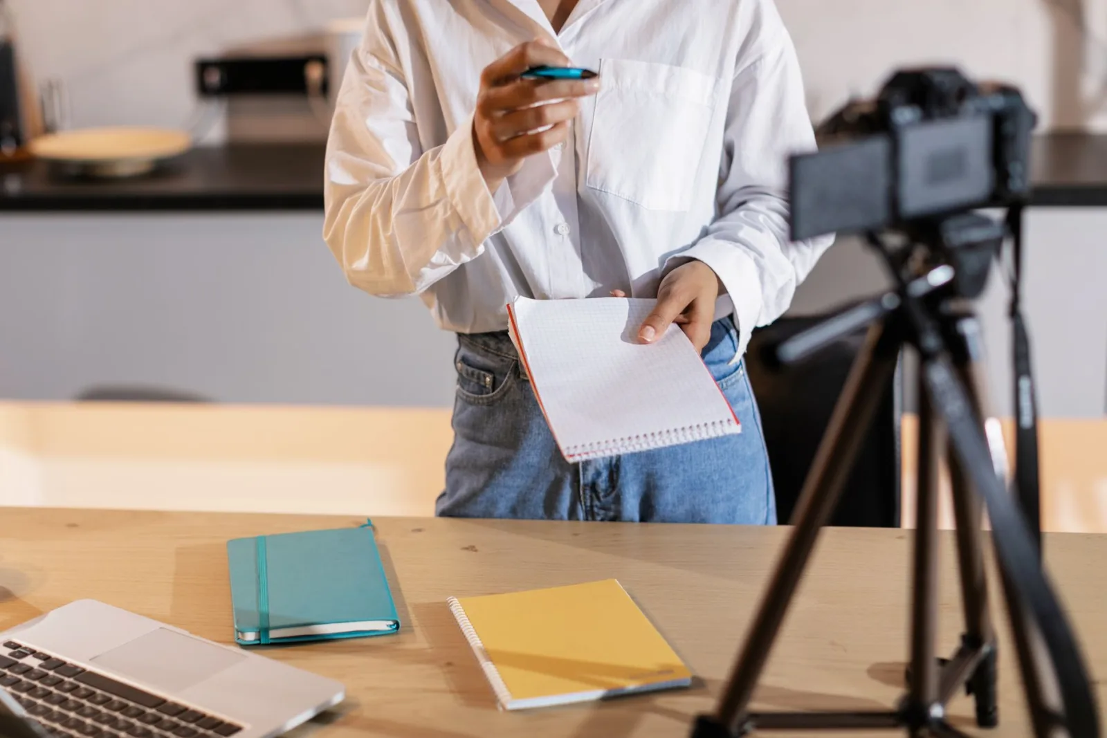 A person in a white shirt holding a spiral notebook and pen beside a camera on a tripod, with notebooks and a laptop on the table.