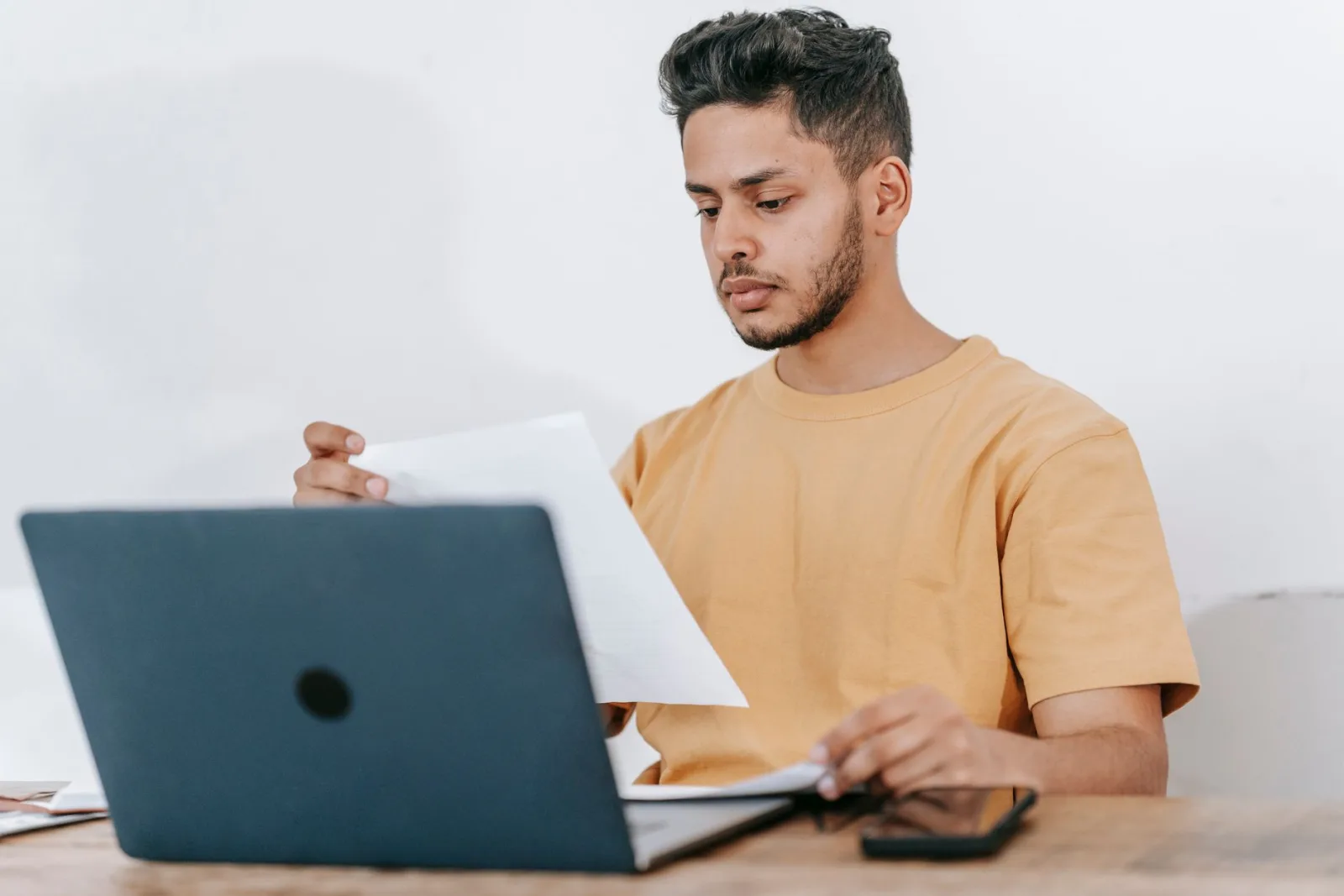 A man in a yellow t-shirt reading a printed document at a wooden desk behind an open laptop.