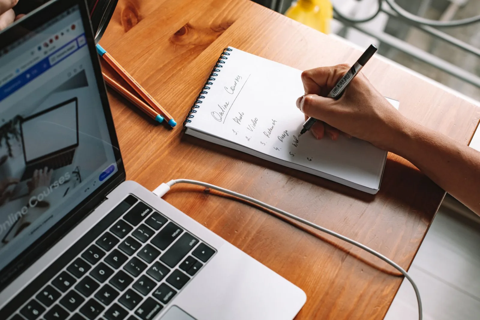 A hand writing a numbered list headed "Online Courses" in a spiral notebook next to an open laptop on a wooden table.